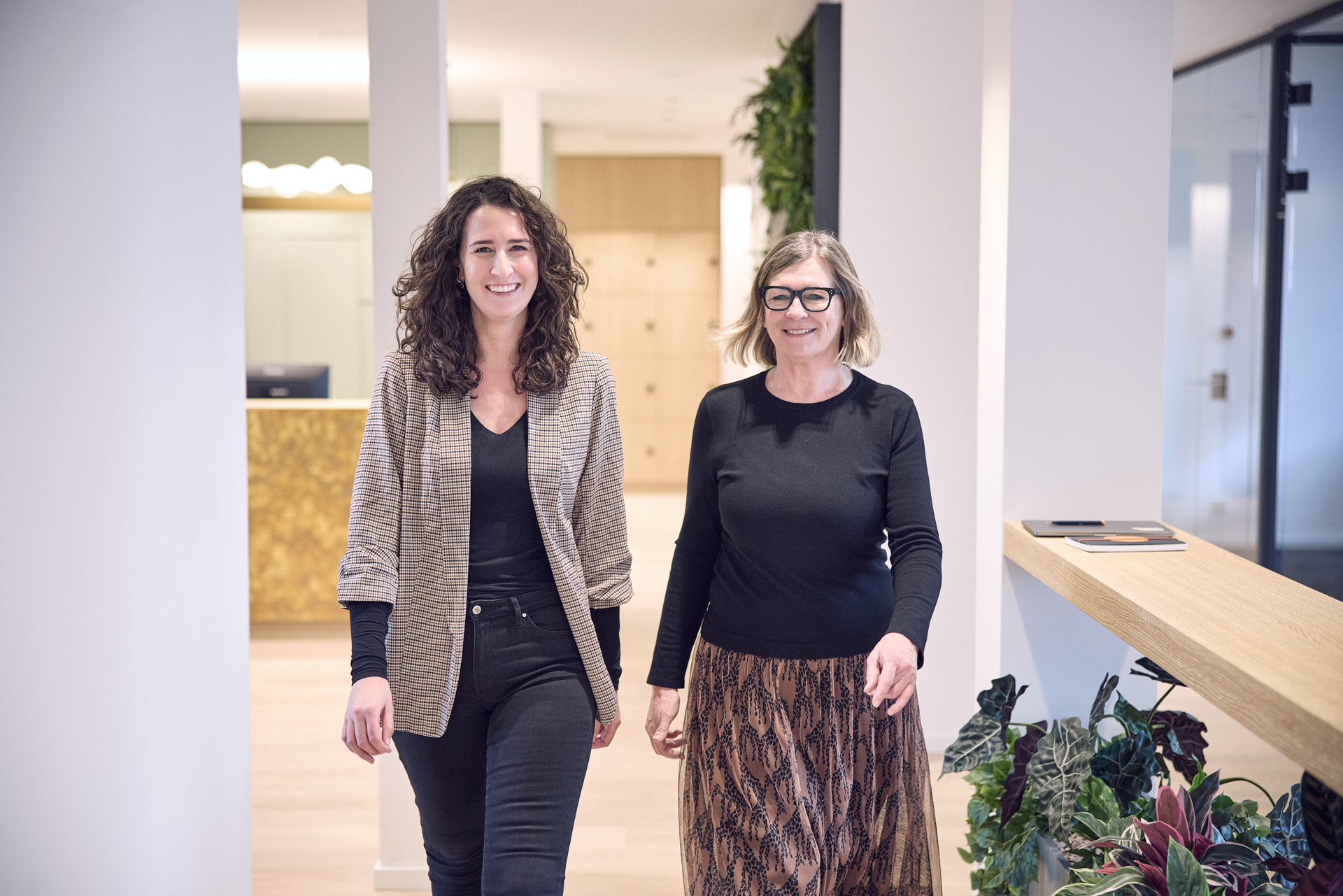 Zwei Frauen gehen lächelnd in einem modernen Bürogebäude einen Gang entlang, umgeben von Pflanzen und hellen Wänden. Die Frau links trägt eine karierte Jacke und schwarze Kleidung, während die Frau rechts ein schwarzes Oberteil und einen gemusterten Rock trägt.