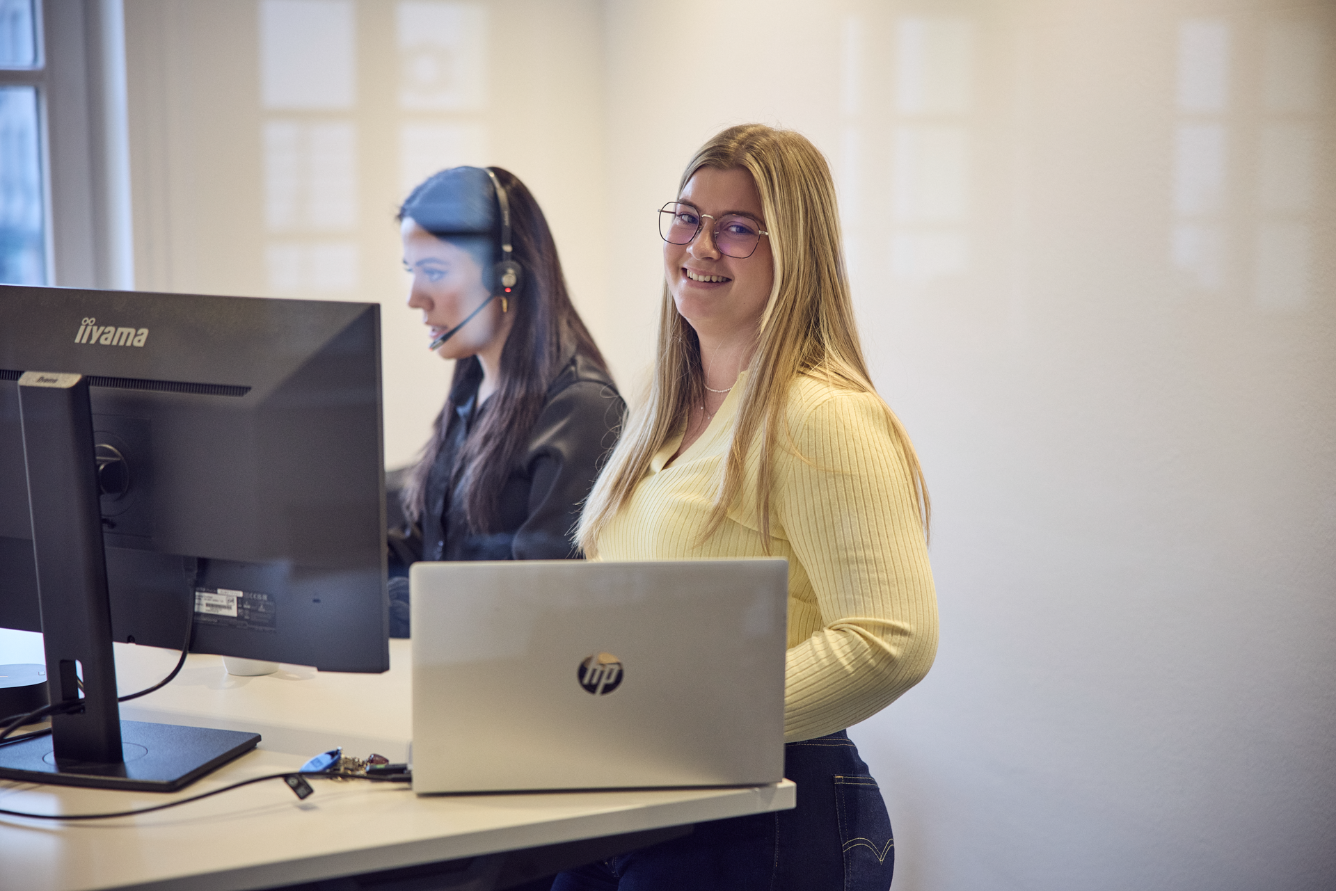 Eine lächelnde Frau mit langen blonden Haaren steht an einem modernen Schreibtisch, während sie in die Kamera schaut; neben ihr sitzt eine weitere Frau mit einem Headset am Computer.