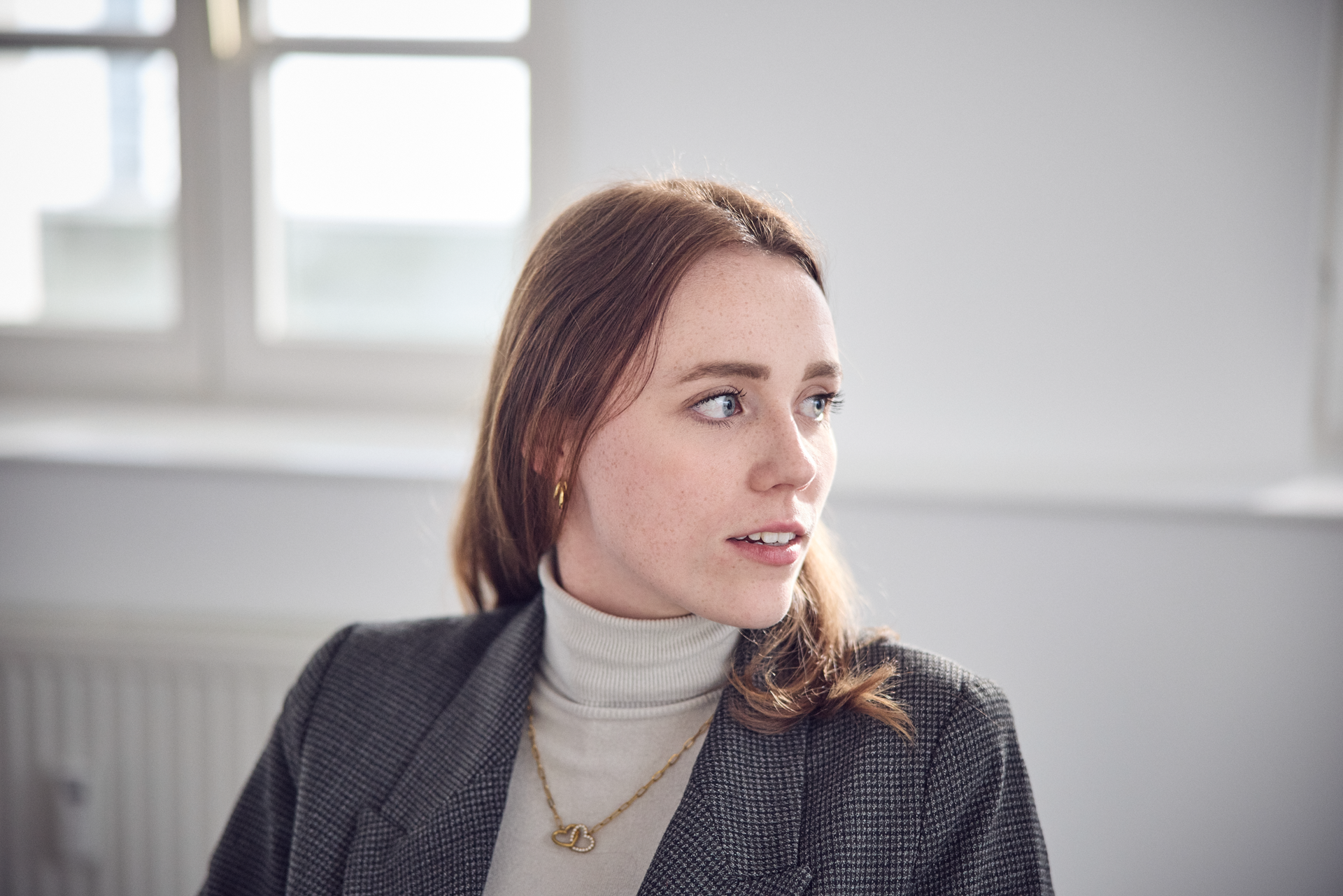 Eine junge Frau mit langen, braunen Haaren trägt einen hohen, grauen Kragenpullover und eine Blazerjacke. Sie sitzt in einem hellen Raum und schaut nachdenklich zur Seite. Ein Fenster im Hintergrund lässt Tageslicht herein.