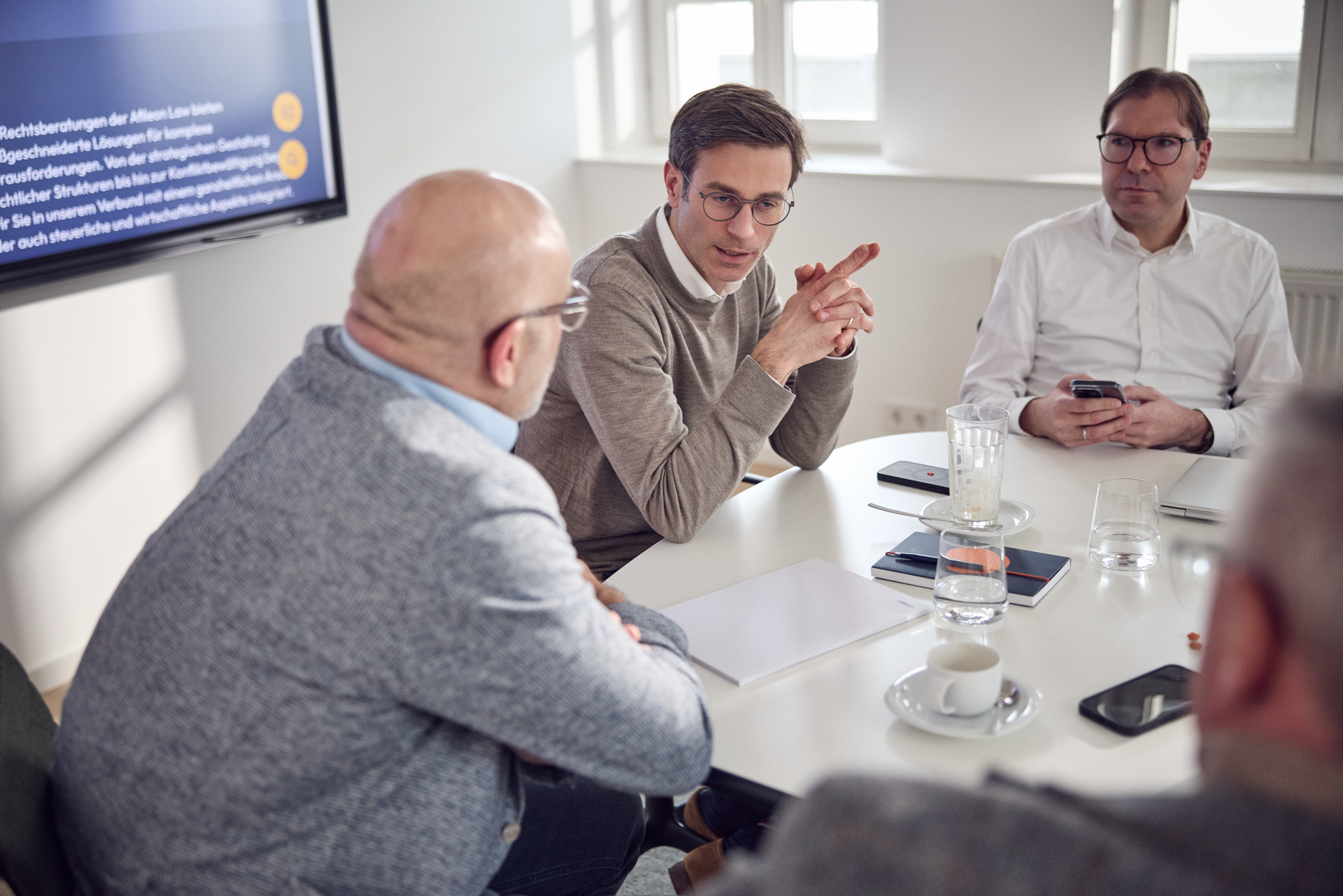 Ein Meeting mit mehreren Teilnehmern an einem Tisch. Ein Mann in einem grauen Pullover spricht anhand von Notizen, während zwei andere Männer ihm zuhören. Auf einem Bildschirm im Hintergrund sind Präsentationsinhalte zu sehen. Auf dem Tisch stehen Gläser und Tassen.