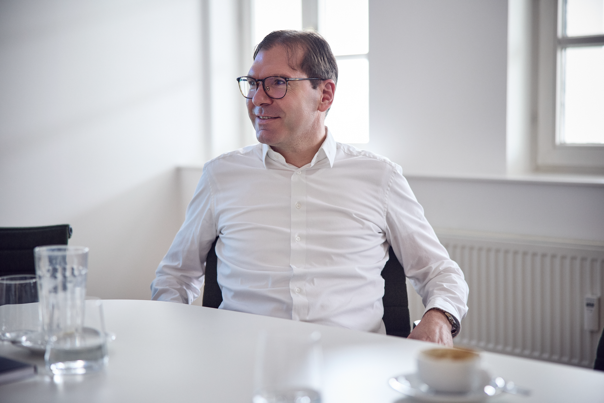 Ein Mann mit Brille sitzt an einem Tisch in einem modernen, hellen Raum und blickt entspannt zur Seite. Auf dem Tisch sind Gläser und eine Kaffeetasse zu sehen.