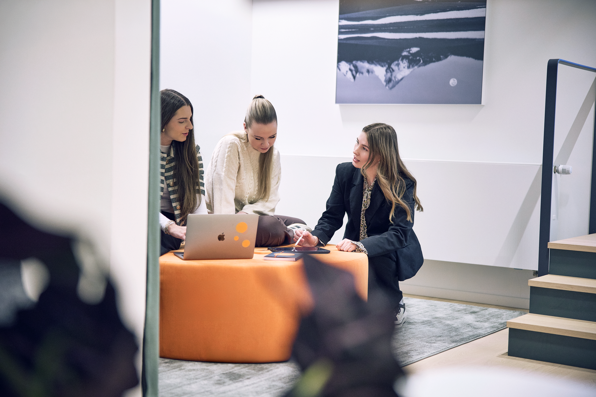 Drei Frauen sitzen gemeinsam auf einem orangen Pouf und schauen auf einen Laptop, während sie an einem Projekt arbeiten. Eine Frau hält ein Handy in der Hand und alle drei scheinen in ein Gespräch vertieft zu sein. Im Hintergrund ist ein abstraktes Bild an der Wand zu sehen.