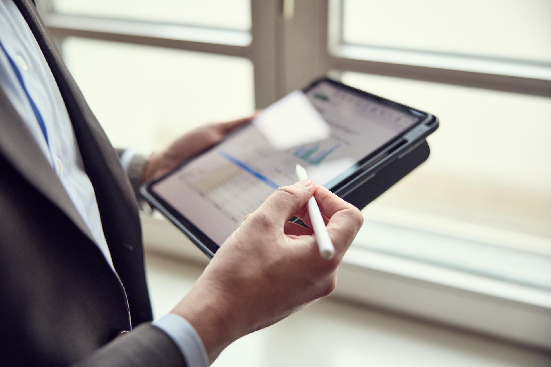 Ein Mann in einem Anzug hält ein Tablet in der Hand und verwendet einen Stylus, während er Daten und Diagramme auf dem Bildschirm betrachtet. Im Hintergrund sind Fenster zu sehen, die Tageslicht hereinlassen.