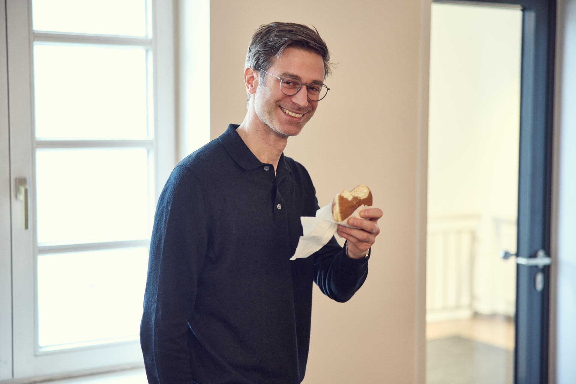Ein Mann mit Brille hält ein belegtes Brötchen in der Hand und lächelt freundlich, während er in einem hellen Raum steht.