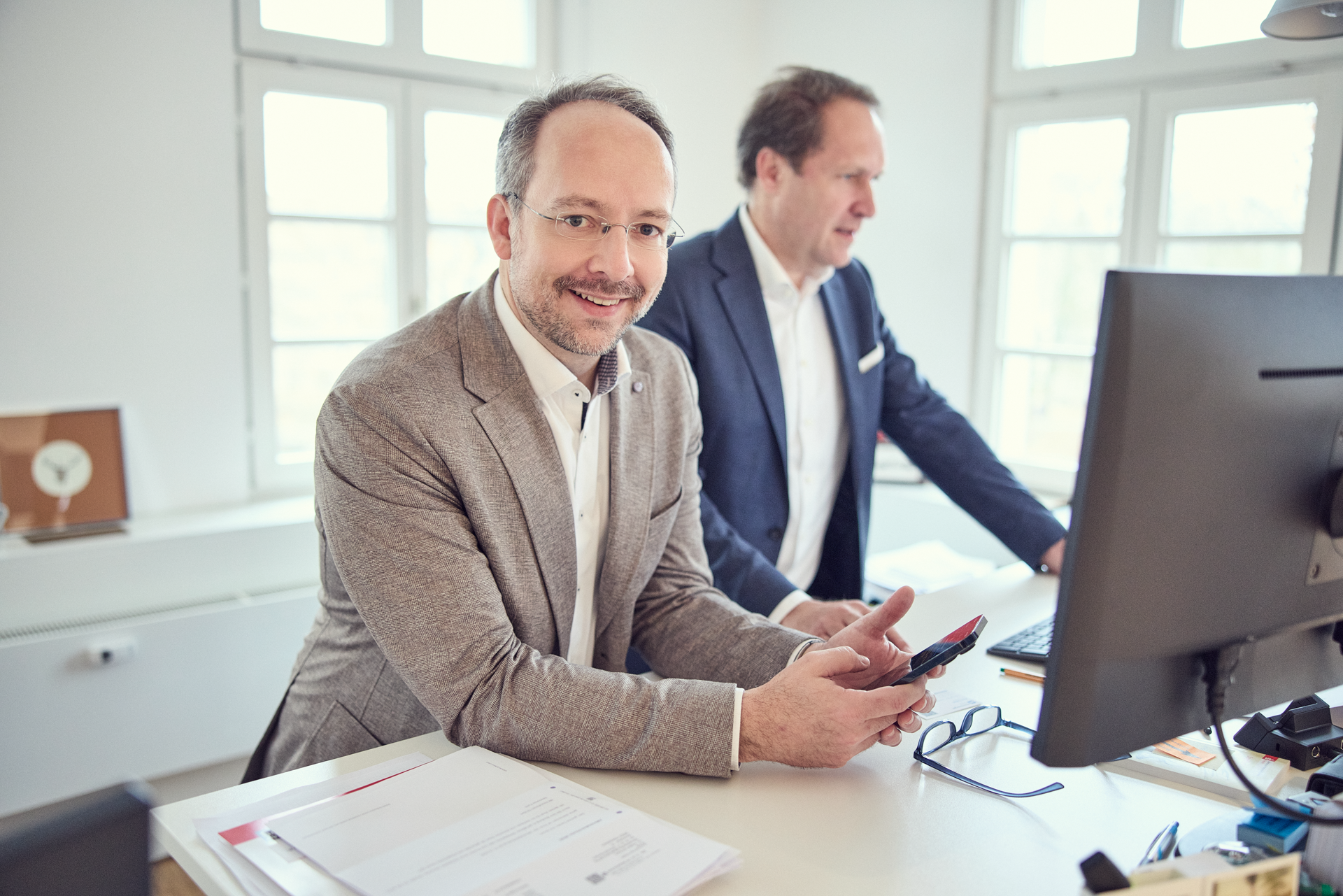 Zwei Männer in einem modernen Büro stehen an einem Tisch; einer von ihnen hält ein Smartphone in der Hand und lächelt in die Kamera, während der andere hinter einem Computerbildschirm steht und auf den Bildschirm schaut. Auf dem Tisch liegen einige Dokumente.