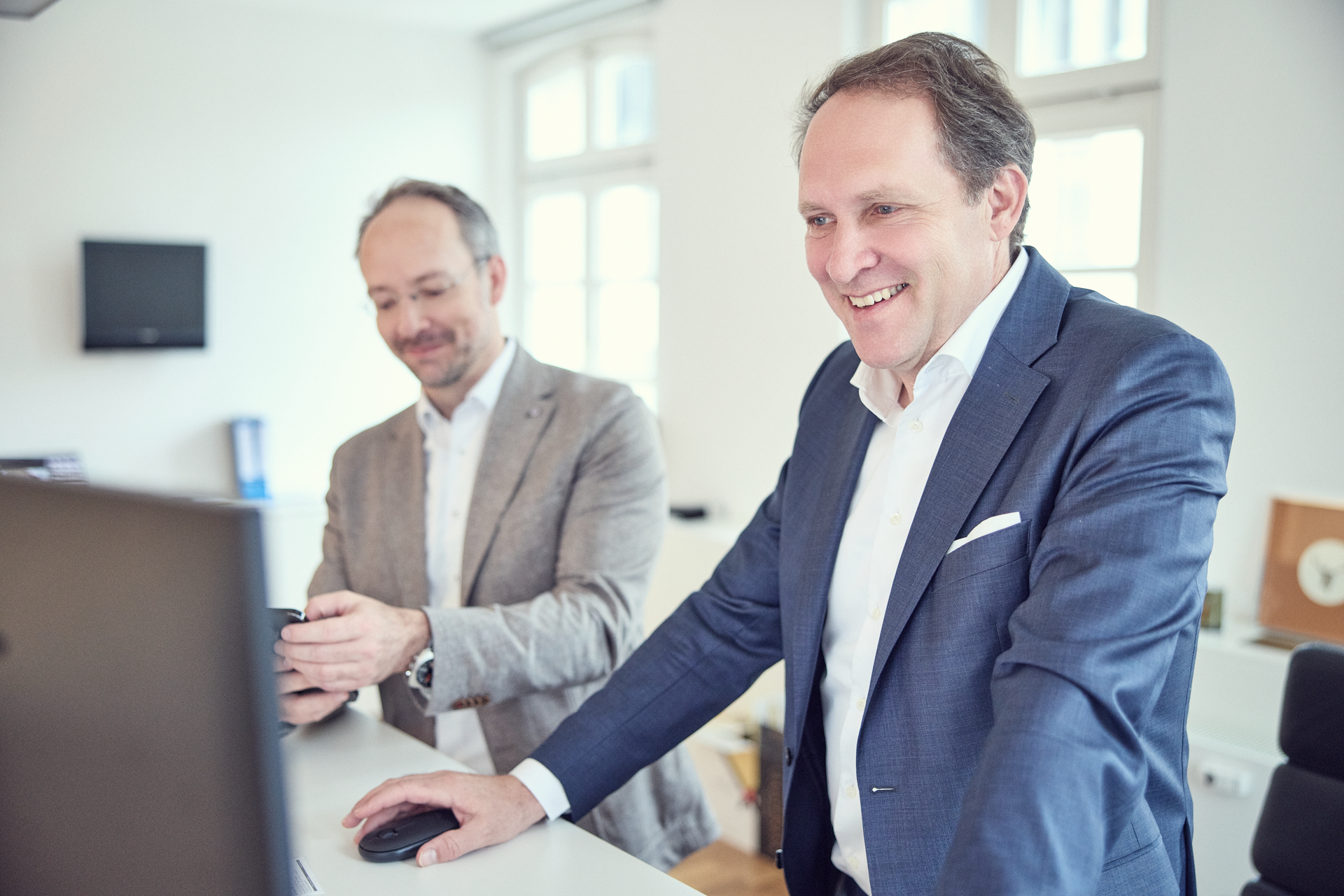 Zwei Männer in formeller Kleidung stehen an einem weißen Schreibtisch; einer von ihnen lächelt und schaut auf einen Computerbildschirm, während der andere mit einem Smartphone in der Hand beschäftigt ist. Im Hintergrund sind Büroelemente wie ein Fernseher und eine Uhr sichtbar.