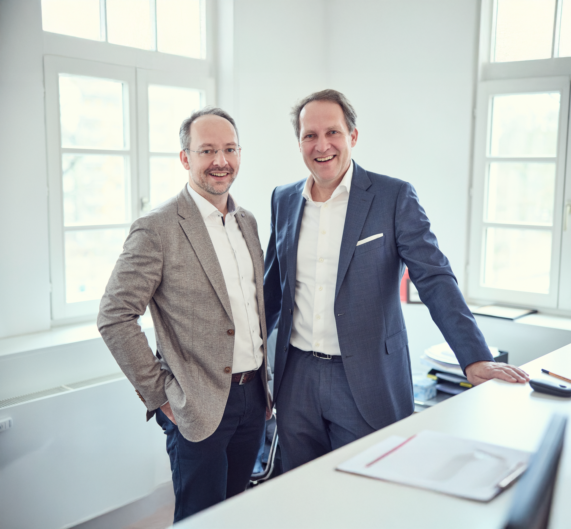 Zwei Männer stehen lächelnd nebeneinander in einem modernen Büroumfeld mit großen Fenstern. Der Mann links trägt einen hellen Anzug, während der Mann rechts einen dunkleren Anzug mit einer weißen Hemdbluse trägt. Auf dem Tisch im Vordergrund sind einige Ordner und Schreibmaterialien zu sehen.