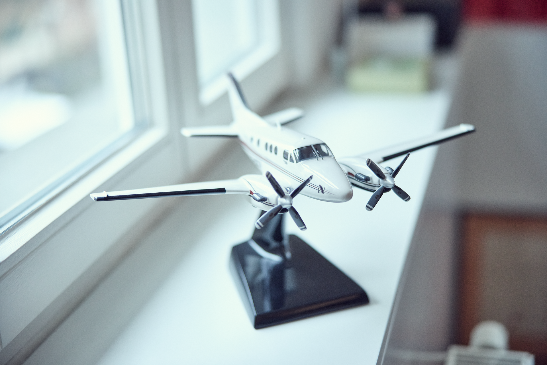 Ein Modellflugzeug mit zwei Propellern steht auf einem Fensterbrett und wird von Tageslicht beleuchtet. Im Hintergrund sind unscharf Möbel und eine Fensterbank zu sehen.