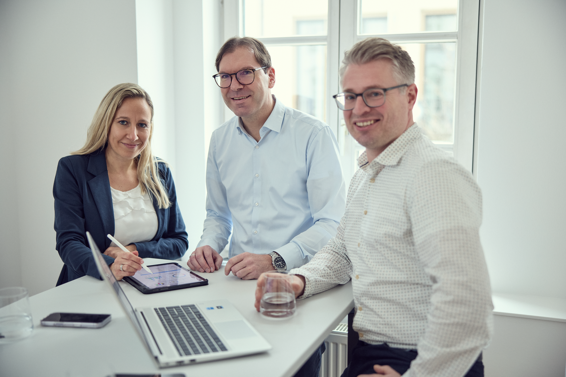 Drei Personen sitzen an einem Tisch in einem hellen, modernen Raum; eine Frau mit langen blonden Haaren und ein Mann in einem hellblauen Hemd verwenden ein Tablet, während ein weiterer Mann mit Brille und einem karierten Hemd lächelt und ein Glas Wasser in der Hand hält.