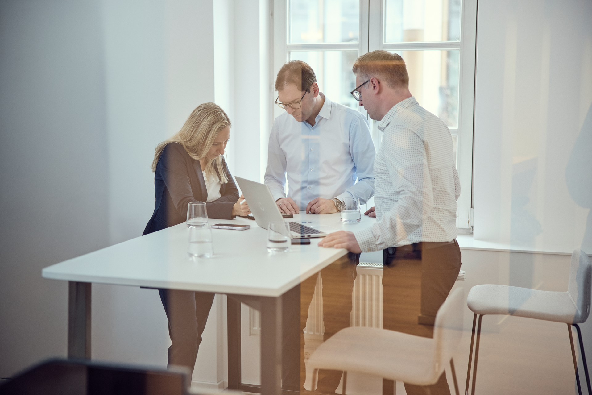 Drei Personen, zwei Männer und eine Frau, stehen an einem Tisch in einem modernen Büro und betrachten einen Laptop; auf dem Tisch stehen Wassergläser.