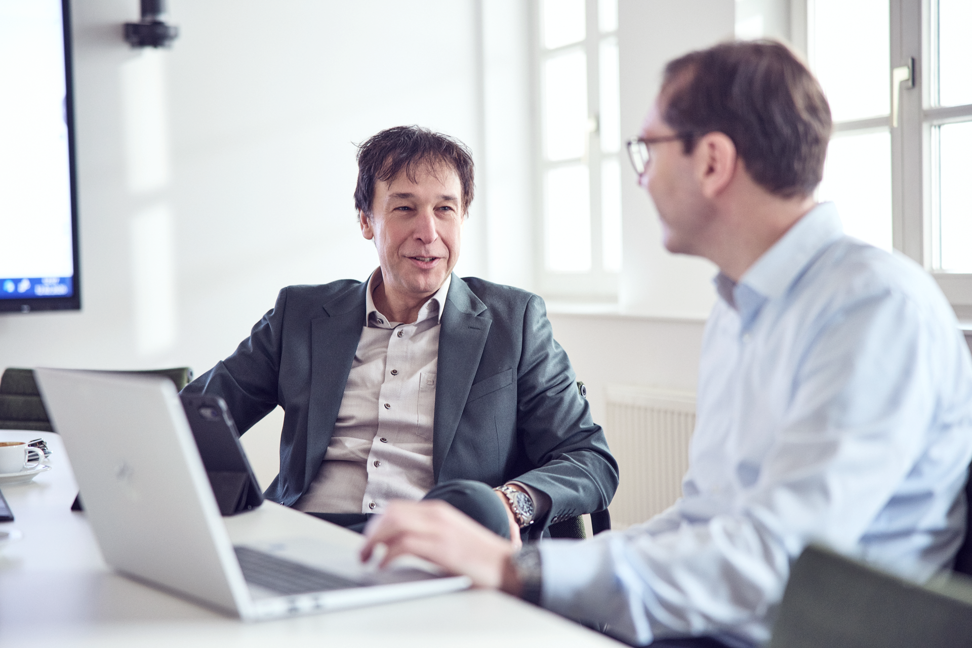 Zwei Männer sitzen an einem Tisch in einem modernen Besprechungsraum und führen ein Gespräch, während ein Laptop und Tassen auf dem Tisch stehen.