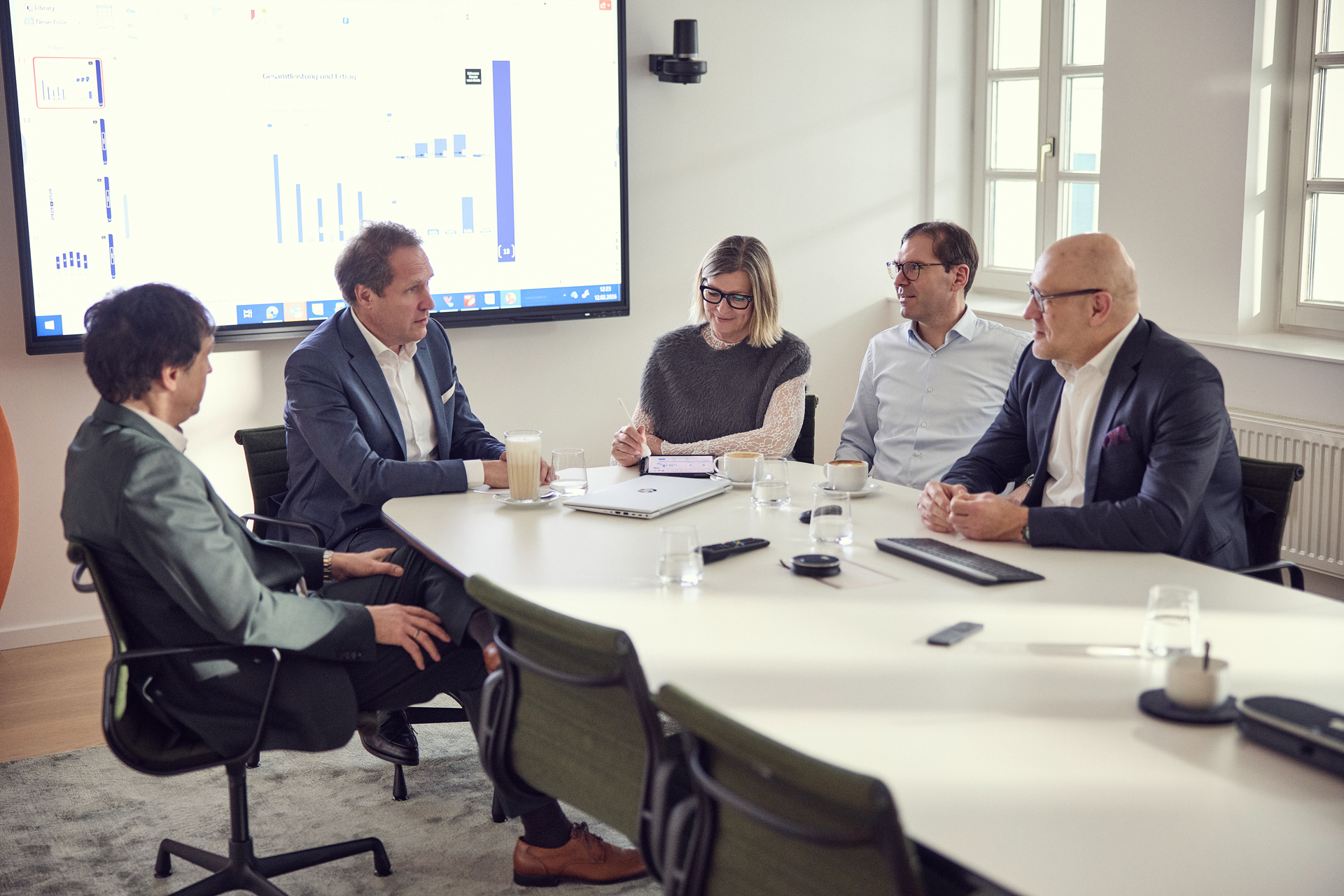 Eine Gruppe von fünf Personen sitzt an einem Konferenztisch in einem modernen Besprechungsraum. Auf einem großen Bildschirm im Hintergrund sind Diagramme und Grafiken zu sehen. Die Teilnehmer sind in Diskussion vertieft, während Notizen gemacht werden.