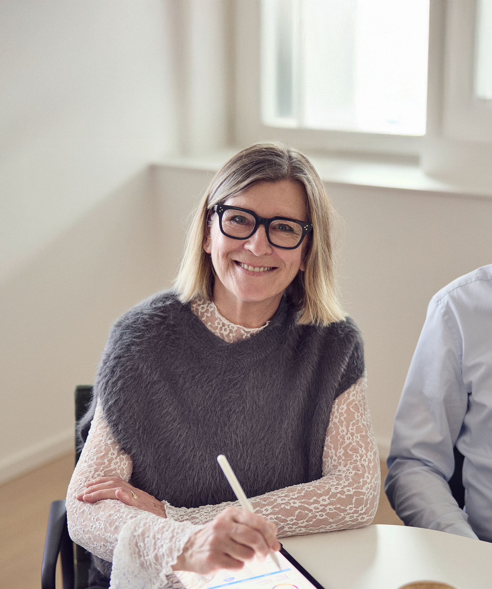 Eine lächelnde Frau mit schulterlangem, blondem Haar trägt ein graues, flauschiges Oberteil über einer weißen, spitzenblumigen Bluse und hält einen Stylus in der Hand, während sie auf einem Tablet zeichnet. Im Hintergrund ist ein heller, moderner Raum mit großen Fenstern zu sehen.