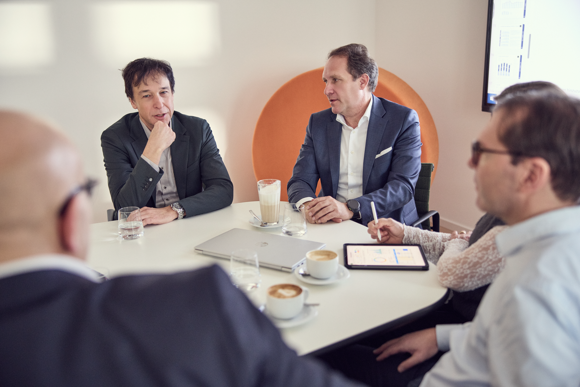 Eine Gruppe von fünf Personen sitzt an einem Tisch in einem Konferenzraum, während sie besprechen und Notizen machen. Zwei der Personen haben Getränke vor sich, und ein Laptop sowie eine Tablette liegen auf dem Tisch. Eine orangefarbene Designerseite ist im Hintergrund sichtbar.