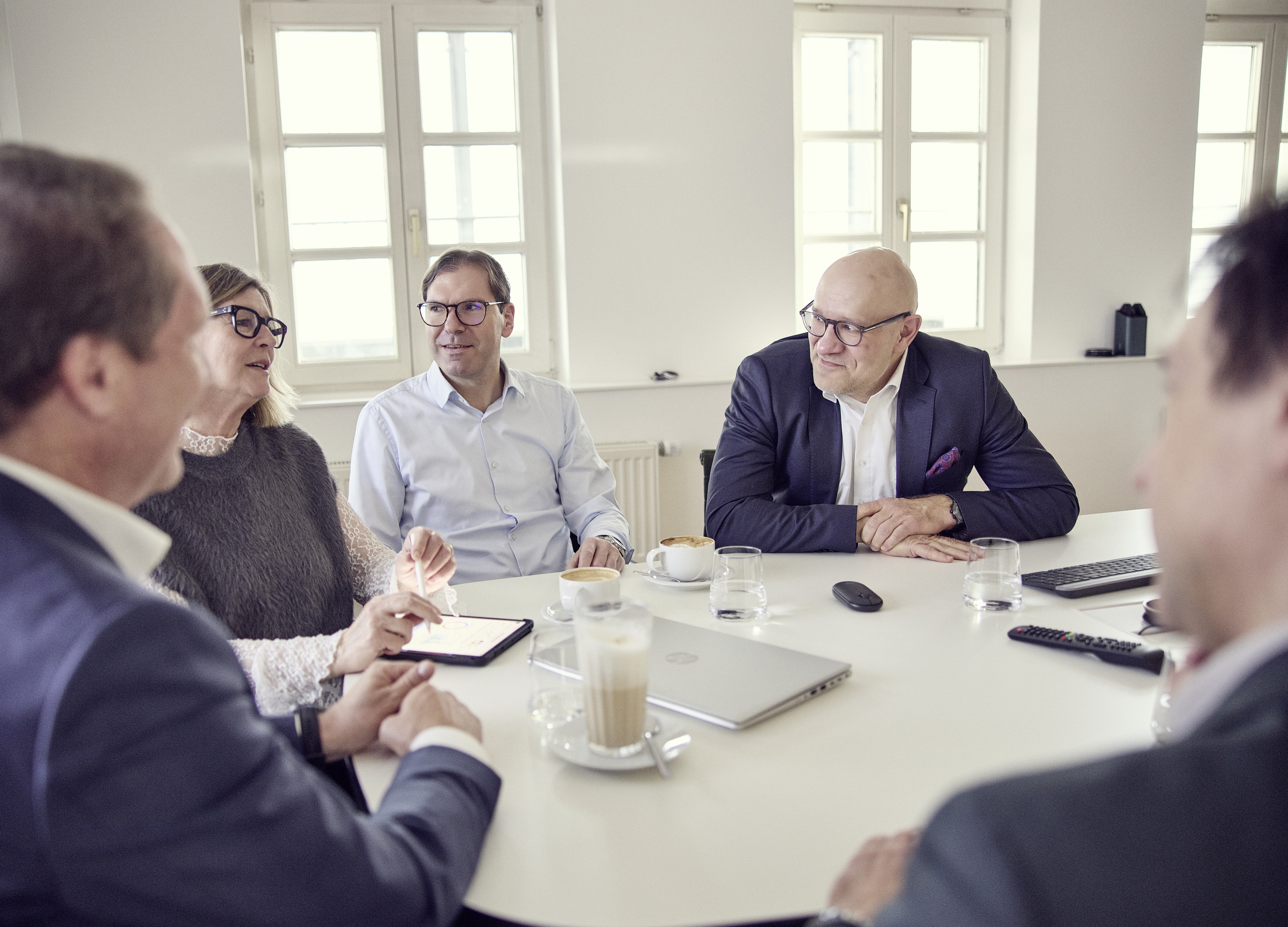 Sechs Personen sitzen um einen Konferenztisch, einige mit Laptops und Getränken, während sie miteinander sprechen und auf ein Tablet schauen. Die Umgebung ist hell mit großen Fenstern.