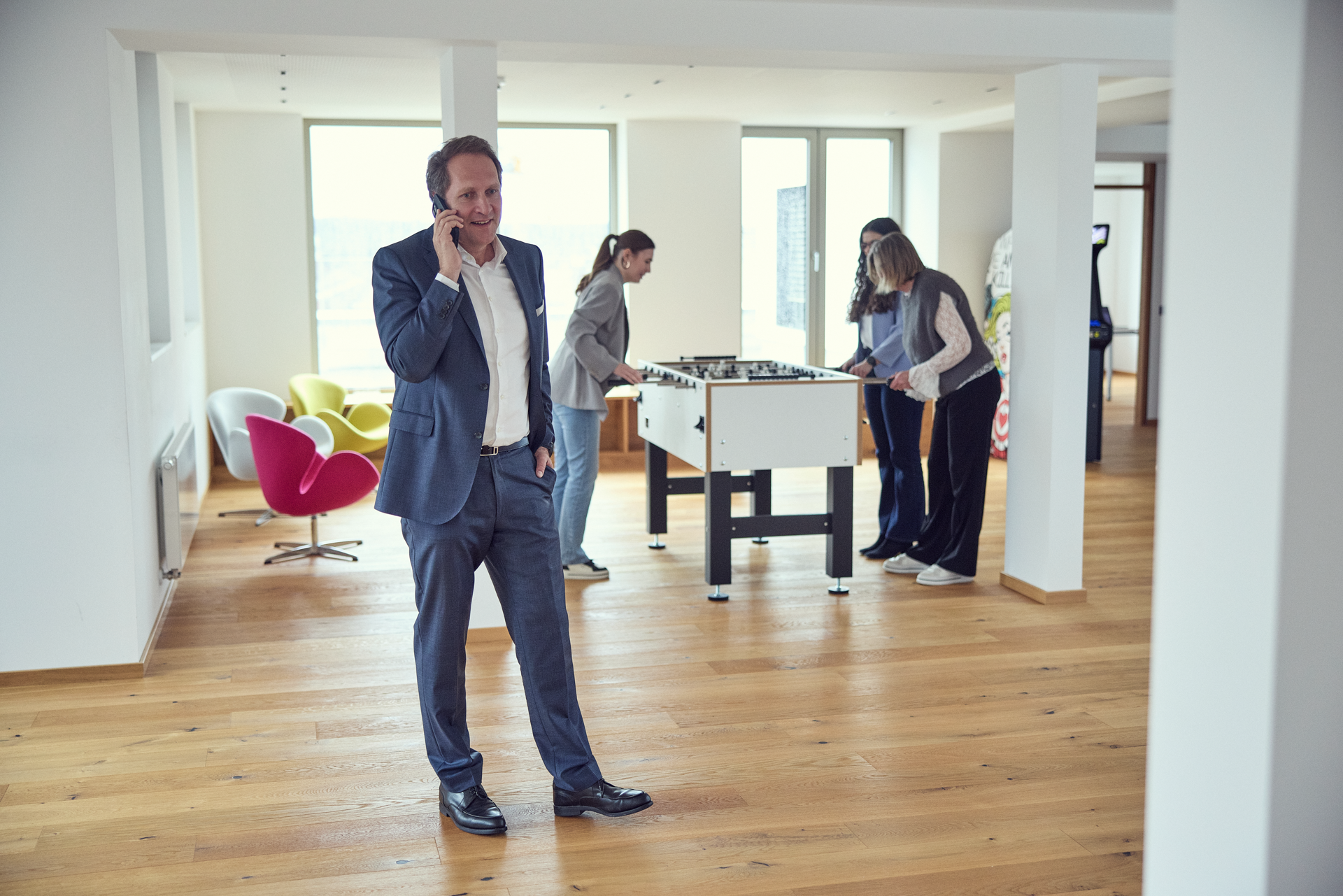 Ein Mann in einem Anzug steht mit einem Handy am Ohr in einem modernen Raum, während im Hintergrund zwei Frauen an einem Tischfußballspiel spielen und eine weitere Person auf einer Couch sitzt. Der Raum hat große Fenster, viel Tageslicht und einen hellen Holzfußboden.