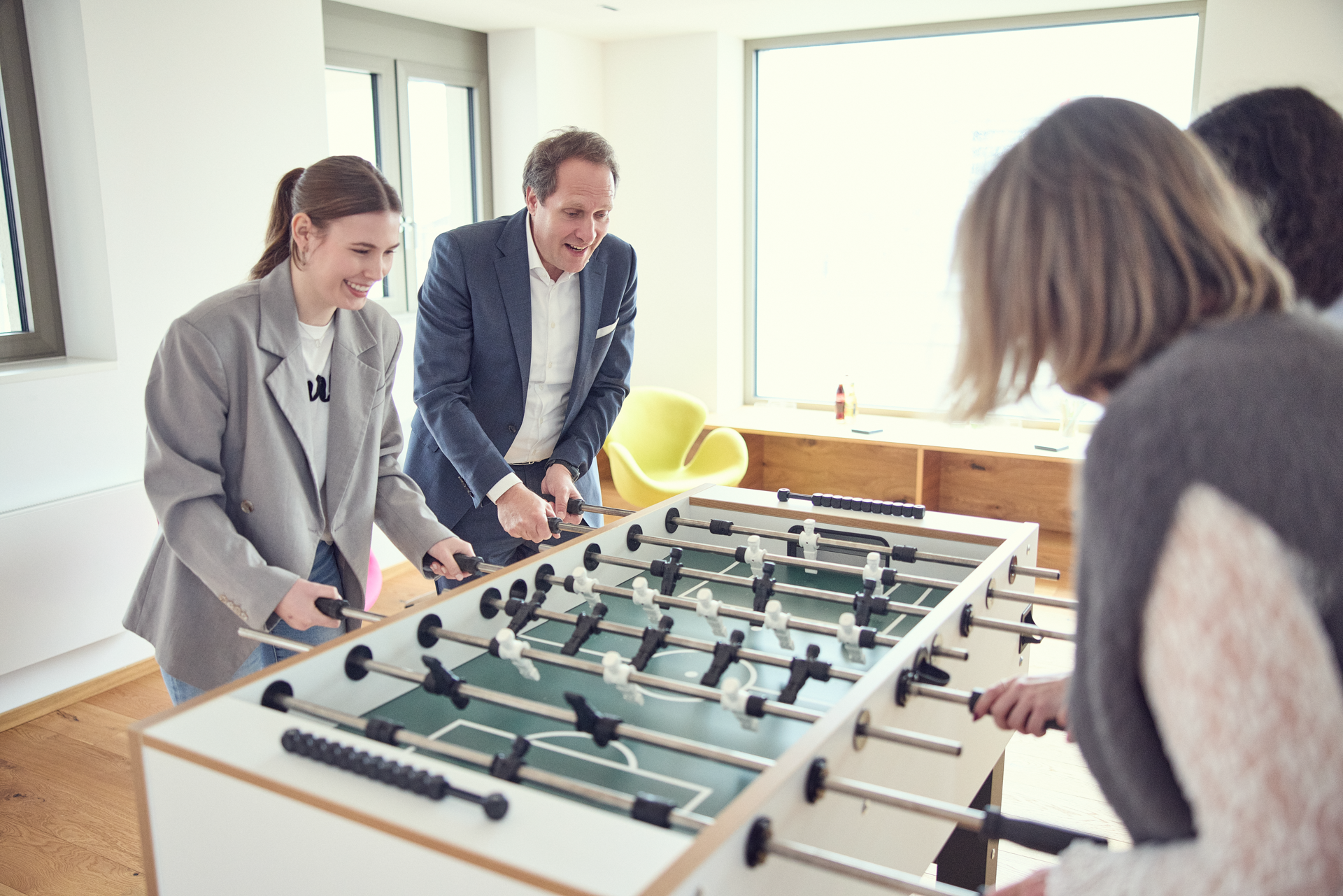 Zwei Personen stehen an einem Tischfußballspiel und spielen gegeneinander, während eine zweite Gruppe zuschaut. Der Mann trägt einen Anzug, die Frau eine große Bluse, und beide scheinen viel Spaß zu haben. Im Hintergrund sind helle Räume mit Fenstern und modernen Möbeln sichtbar.