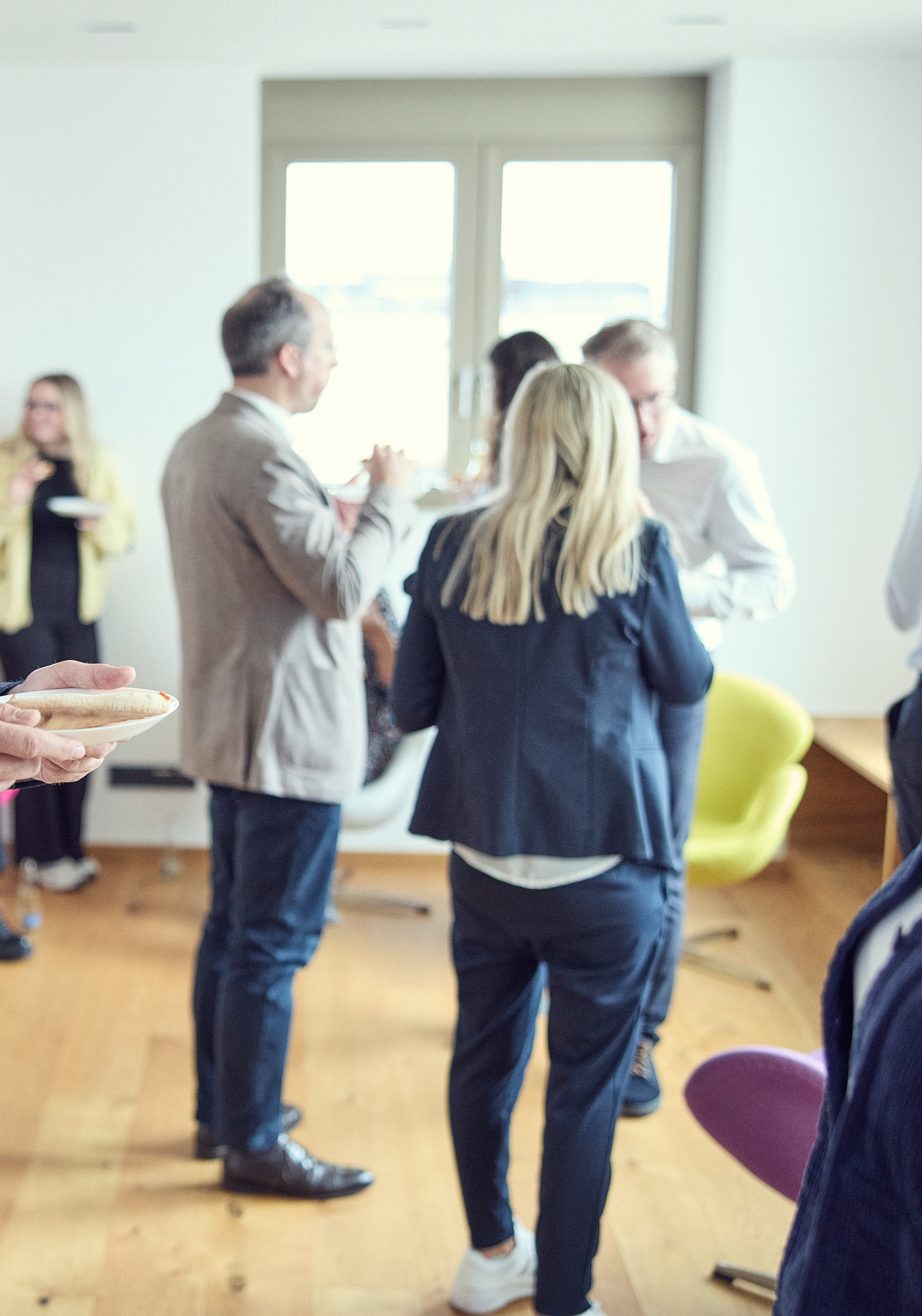 Eine Gruppe von Personen steht in einem hellen Raum, einige halten Teller mit Snacks, während andere miteinander sprechen. Im Vordergrund ist eine Person mit blonden Haaren und einem dunkelblauen Anzug zu sehen, die von hinten gezeigt wird. Im Hintergrund sind weitere Gäste erkennbar.