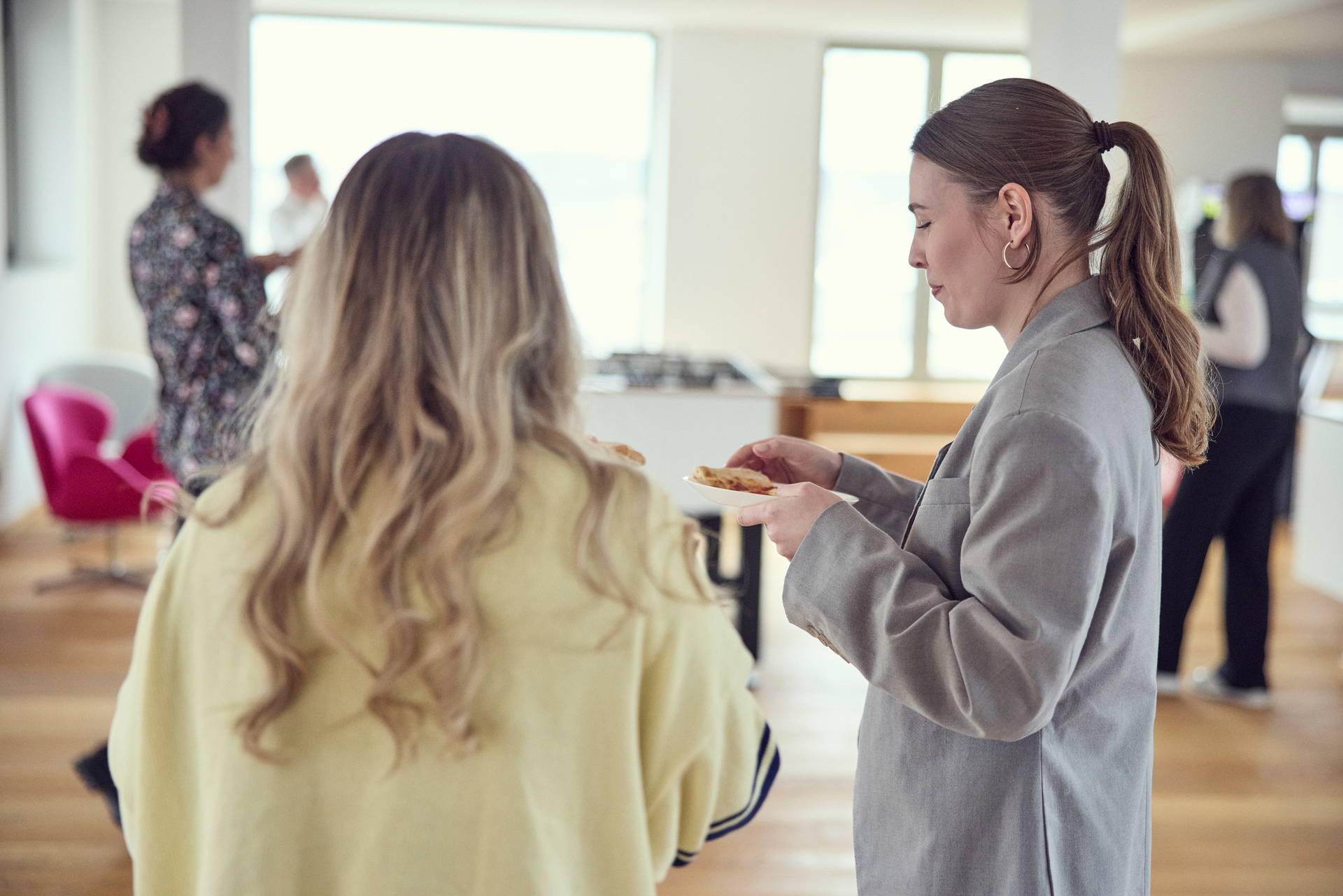 Zwei Frauen stehen in einer modernen, hellen Büroumgebung und unterhalten sich, während sie Snacks auf Tellern halten. Im Hintergrund sind weitere Personen zu sehen, die sich in der lockeren Atmosphäre bewegen.