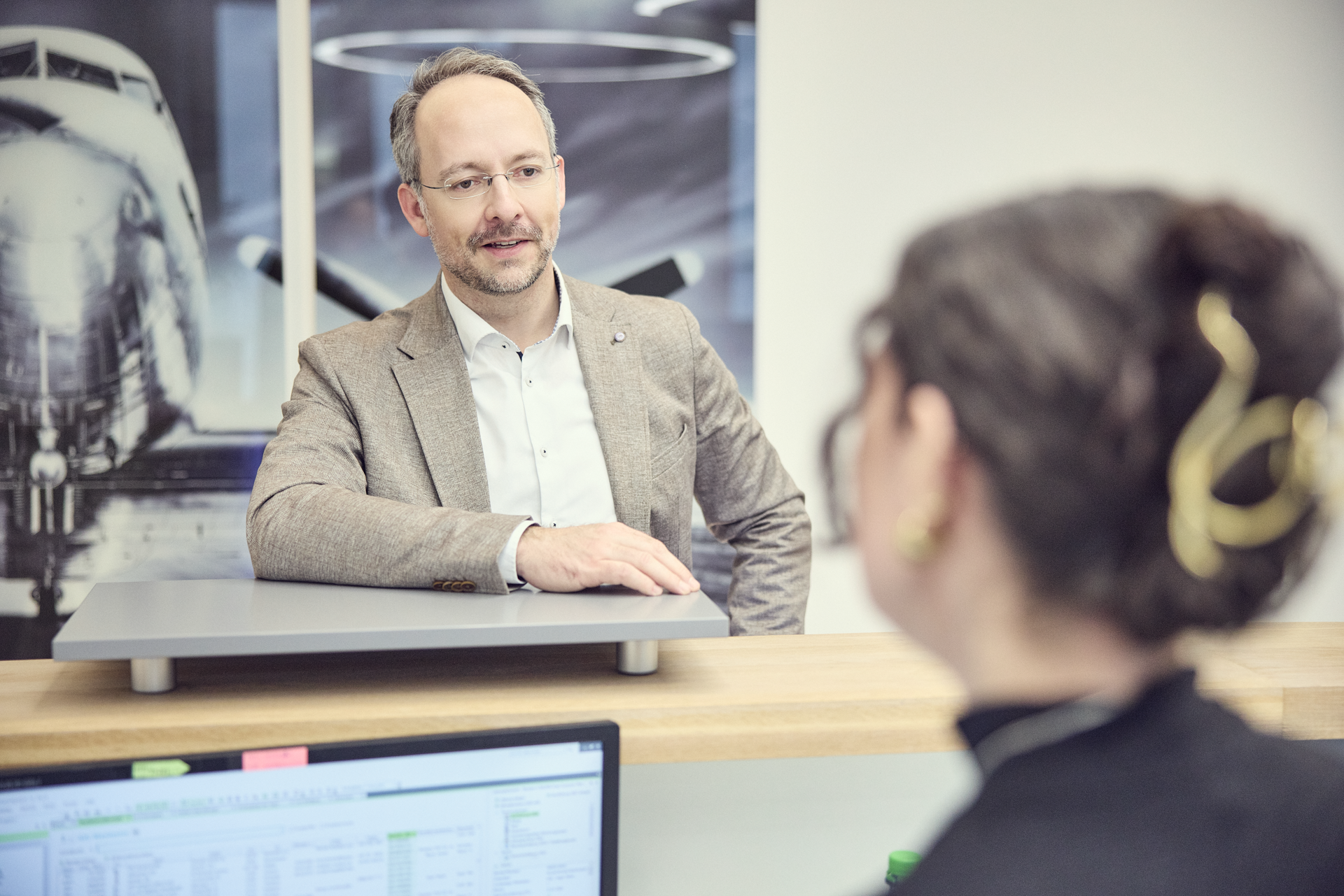 Ein Mann in einem hellen Sakko steht an einem Empfangstresen und unterhält sich mit einer Frau, die ihm den Rücken zugewandt hat. Im Hintergrund ist ein Bild eines Flugzeugs zu sehen.