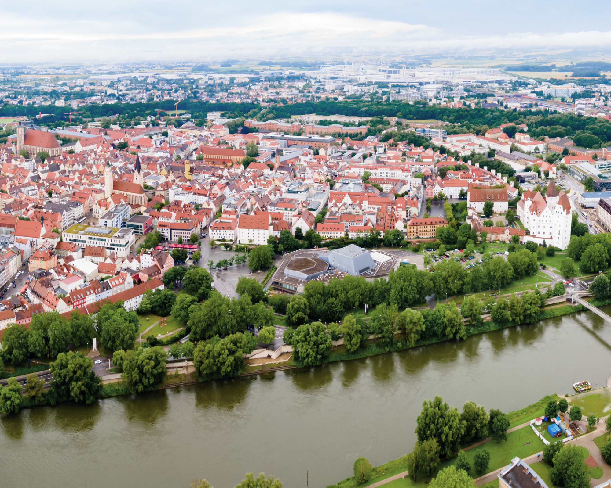 Luftaufnahme einer Stadt mit roten Dächern, umgeben von grünen Bäumen und einem Fluss, der sich durch die Landschaft schlängelt. Im Hintergrund sind moderne Gebäude und eine weite Landschaft zu sehen.