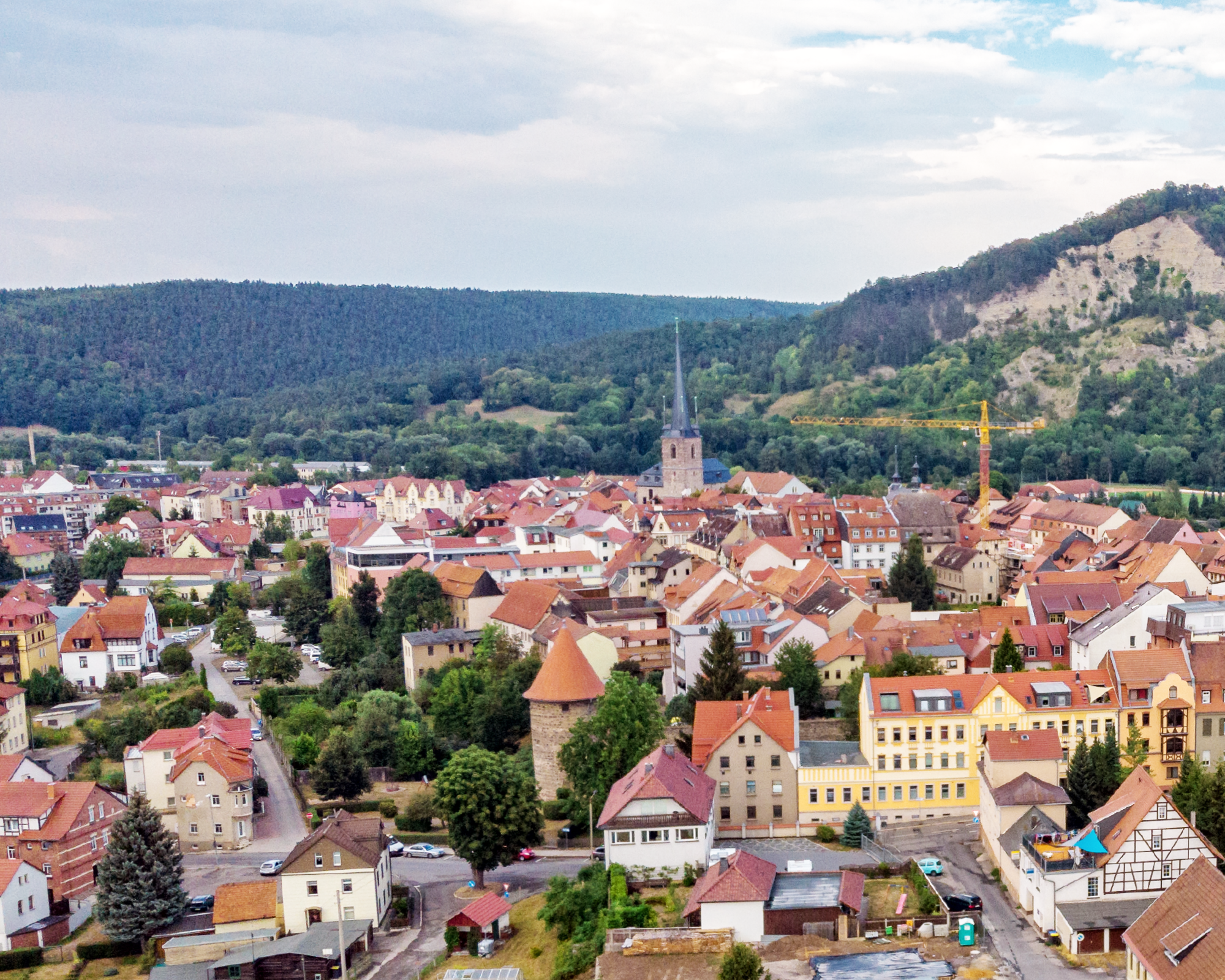 Panoramaansicht einer kleinen Stadt mit roten Dachziegeln, umgeben von bewaldeten Hügeln und einer Kirche im Zentrum. Der Himmel ist teilweise bewölkt und im Hintergrund ist eine Baustelle mit einem Kran zu sehen.