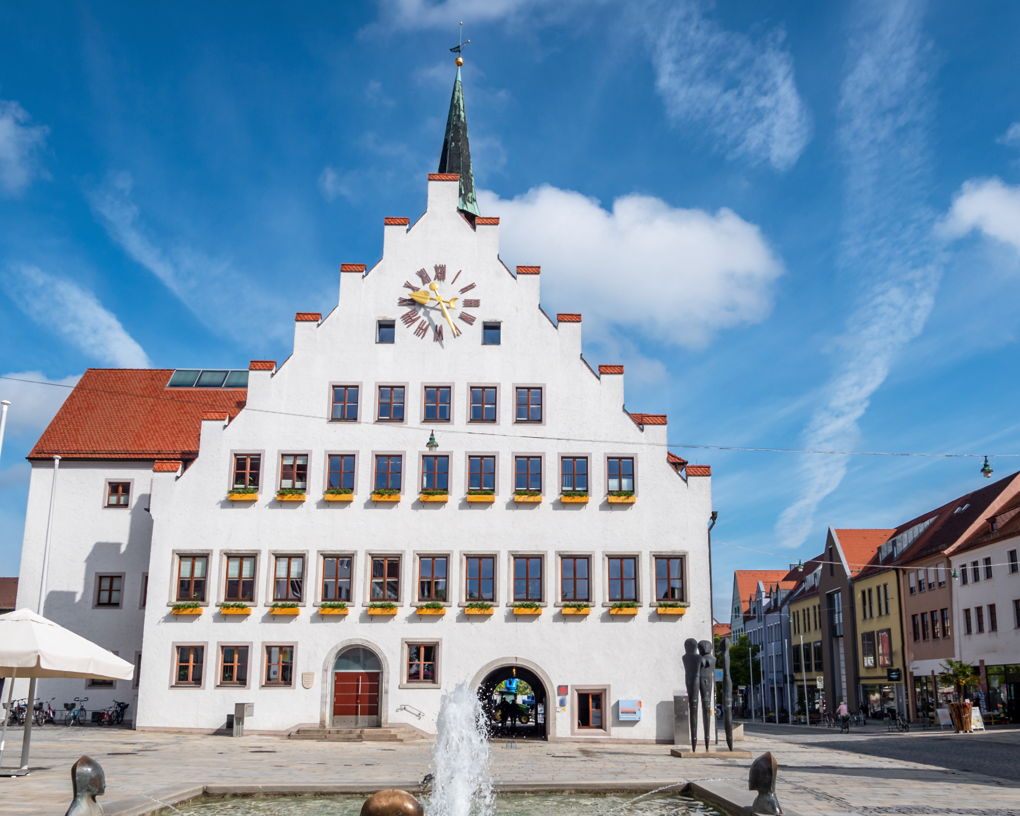 Das Rathaus mit einem markanten, weiß getünchten Giebel und einem Ziffernblatt an der Fassade steht auf einem Platz, umgeben von weiteren Gebäuden. Ein Brunnen mit Wasserstrahl befindet sich im Vordergrund, und der Himmel ist klar mit einigen Wolken.