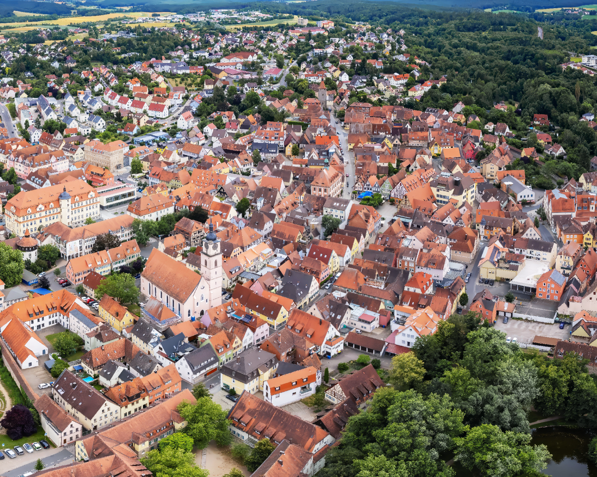 Eine Luftaufnahme einer malerischen Stadt mit zahlreichen roten Dächern und einer historischen Kirche im Zentrum, umgeben von grünen Wäldern und Feldern.