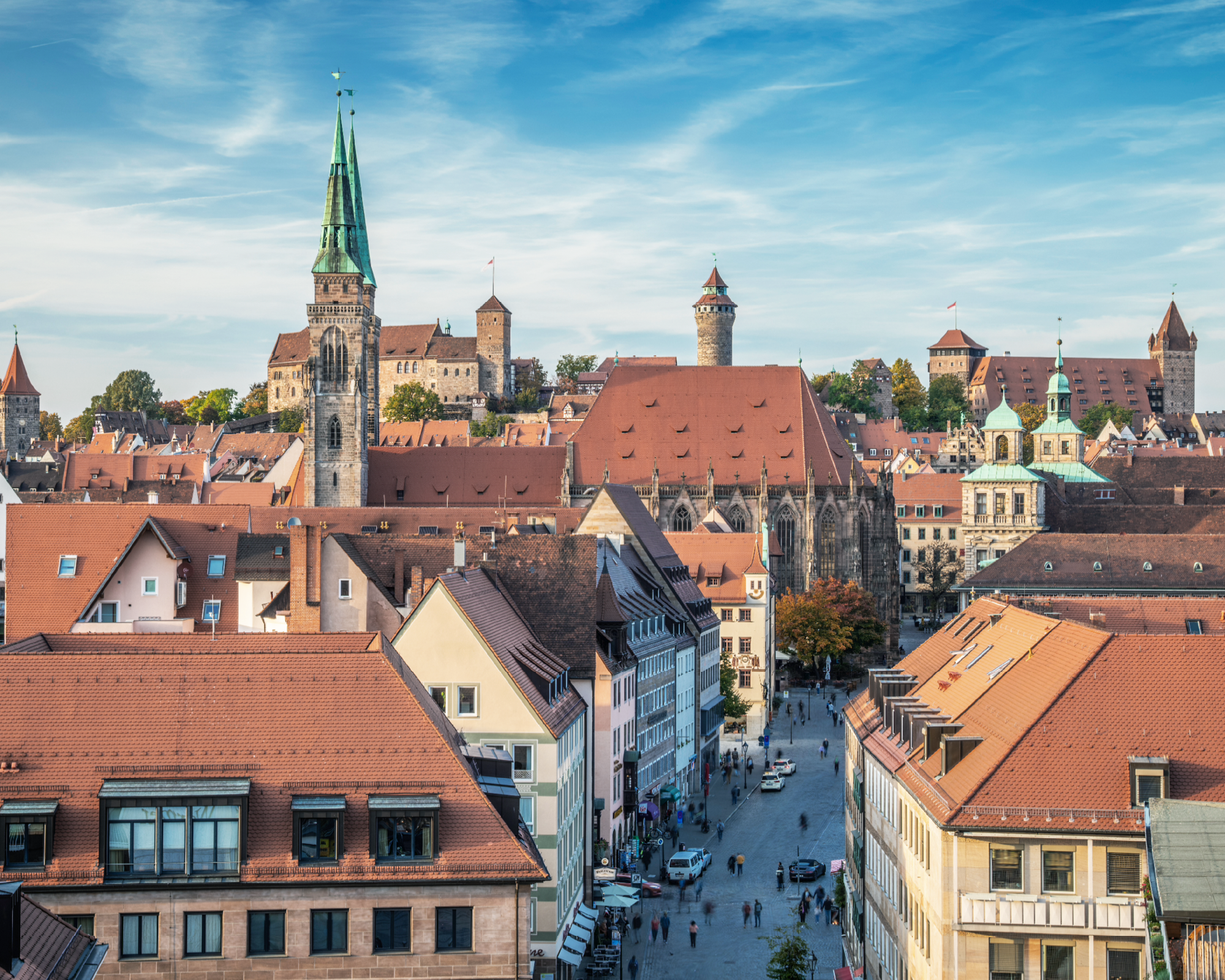 Blick auf die Altstadt von Nürnberg, mit roten Ziegeldächern und historischen Gebäuden, darunter der Hauptmarkt und die Kirche St. Sebald, umgeben von Hügeln und einem klaren blauen Himmel.