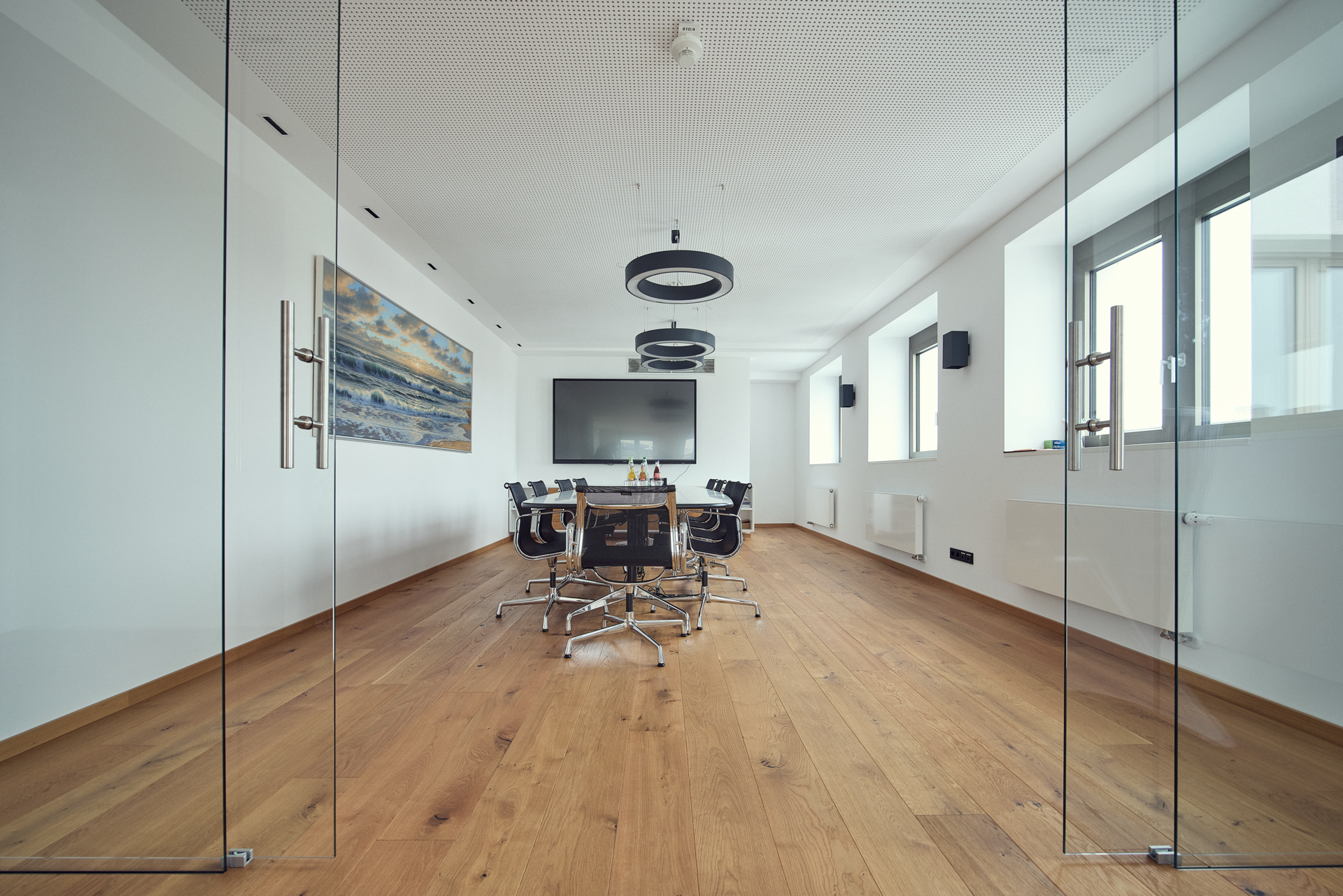 Ein moderner Konferenzraum mit Holzboden, einem großen Tisch in der Mitte, umgeben von Drehstühlen, und einem Bildschirm an der Wand. Große Fenster lassen viel Tageslicht herein, und an einer Wand hängt ein Bild mit einer Landschaft.