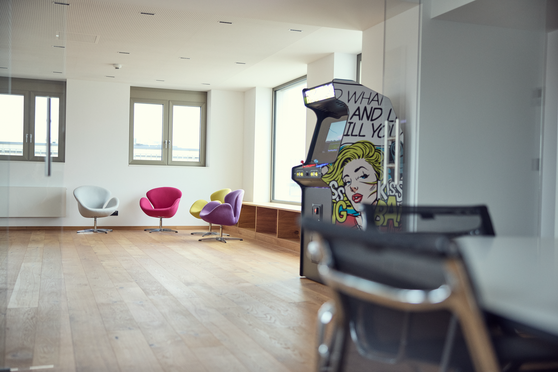 Ein moderner Büroraum mit hellen Wänden und großen Fenstern, in dem ein stilvoller Tisch mit Stühlen, ein paar farbige Sitzmöbel in Pink und Weiß sowie ein Retro-Spiel-Automat mit einem bunten Comic-Design stehen.