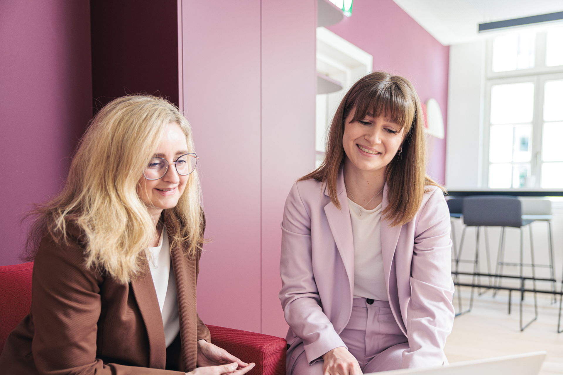 Zwei Frauen sitzen auf einem roten Sofa in einem modernen Büro und betrachten gemeinsam einen Laptop. Eine Frau trägt eine braune Blazerjacke und hat lockiges Haar, während die andere eine hellrosafarbene Anzugjacke trägt und glattes Haar hat. Beide lächeln und wirken konzentriert. Im Hintergrund sind sichtbare Büromöbel und eine Fensterfront.
