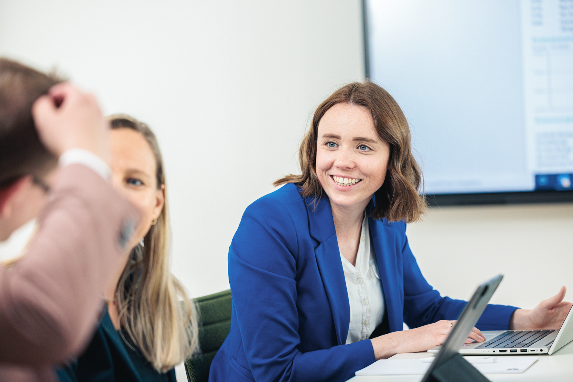 Eine lächelnde Frau mit braunen Haaren trägt einen blauen Blazer und sitzt an einem Tisch in einem modernen Konferenzraum, während sie aktiv an einer Besprechung teilnimmt. Im Hintergrund sind zwei weitere Personen zu sehen, die ebenfalls in die Diskussion eingebunden sind.