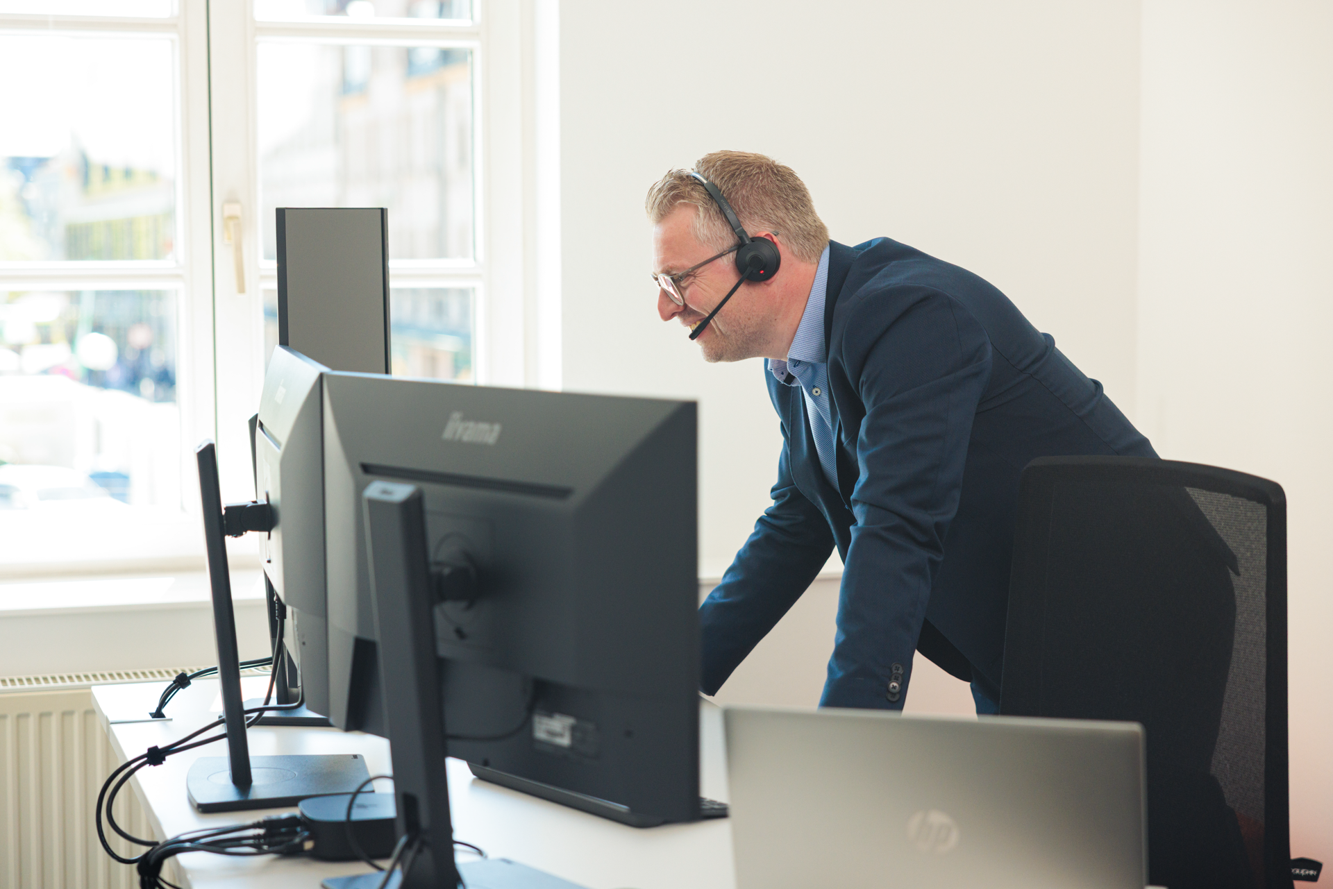 Ein Mann steht an einem Schreibtisch mit mehreren Monitoren und trägt ein Headset. Er scheint in ein Gespräch vertieft zu sein und schaut auf die Bildschirme.